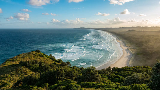 Golden sunrise over Byron Bay's sweeping coastline, with rolling waves, white sand beach, and lush green headland in the foreground.