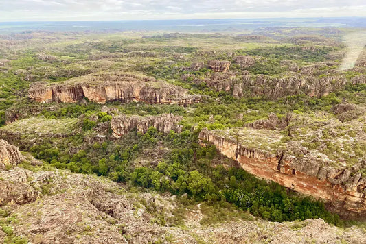 Aerial view of Kakadu National Park showing sandstone escarpments, red rock formations, and dense green bushland stretching to the horizon.